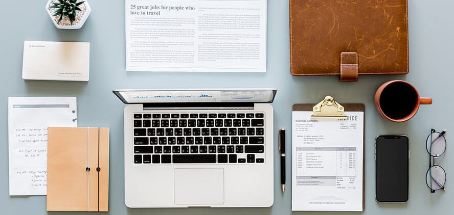 The image shows a neatly organized workspace with a laptop, documents, a notebook, a planner, a coffee cup, a smartphone, and office supplies