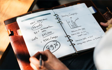 a person writing in a notebook or planner with business-related notes, including numbers, percentages, and a graph
